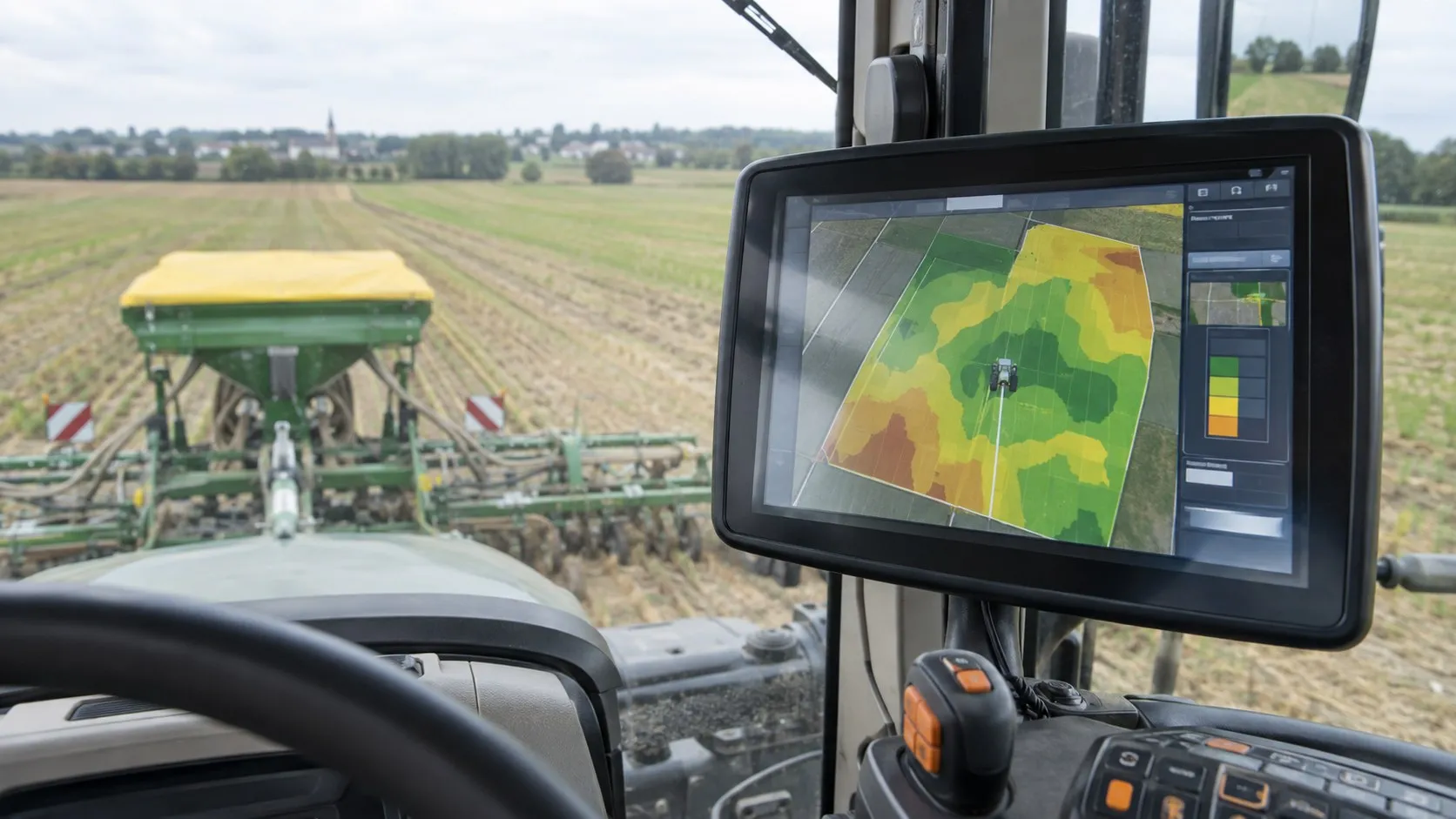 Console de guidage dans la cabine d’un tracteur affichant une carte de modulation en zones colorées, l’épandeur au travail visible par la vitre.